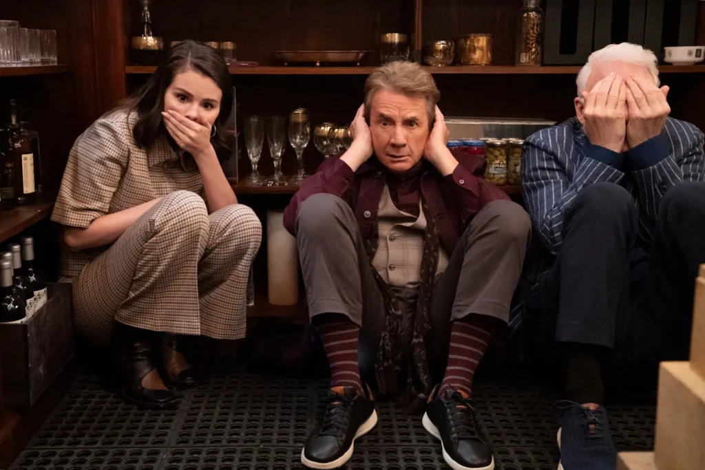 Trois personnes sont assises par terre dans un débarras ; l'une se couvre la bouche, l'autre les oreilles et la troisième le visage, comme une scène tout droit sortie de Only Murders In The Building Saison 5, actuellement en streaming sur Disney+.