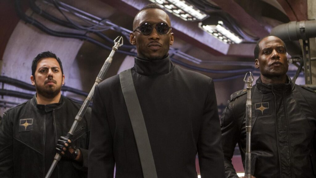 Trois hommes en uniforme noir et lunettes de soleil se tiennent à l'intérieur ; la figure centrale, inspirée par Marvel et Mahershala Ali, porte une ceinture tandis que les deux autres tiennent des lances.