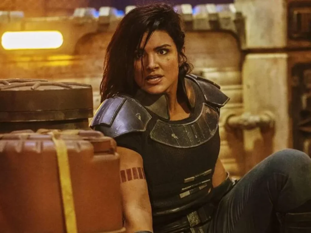 Une femme en armure de science-fiction, rappelant les rôles de Gina Carano, est assise sur le sol dans un environnement industriel, l'air alerte et concentré.