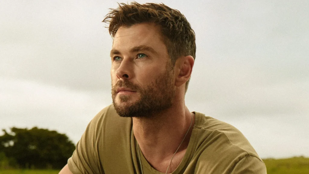 Un homme aux cheveux bruns courts et à la barbe taillée regarde au loin en plein air, vêtu d'un t-shirt kaki et d'un collier - son style rappelle celui de Chris Hemsworth dans Limitless de Disney+. Des arbres et un ciel nuageux sont à l'arrière-plan.