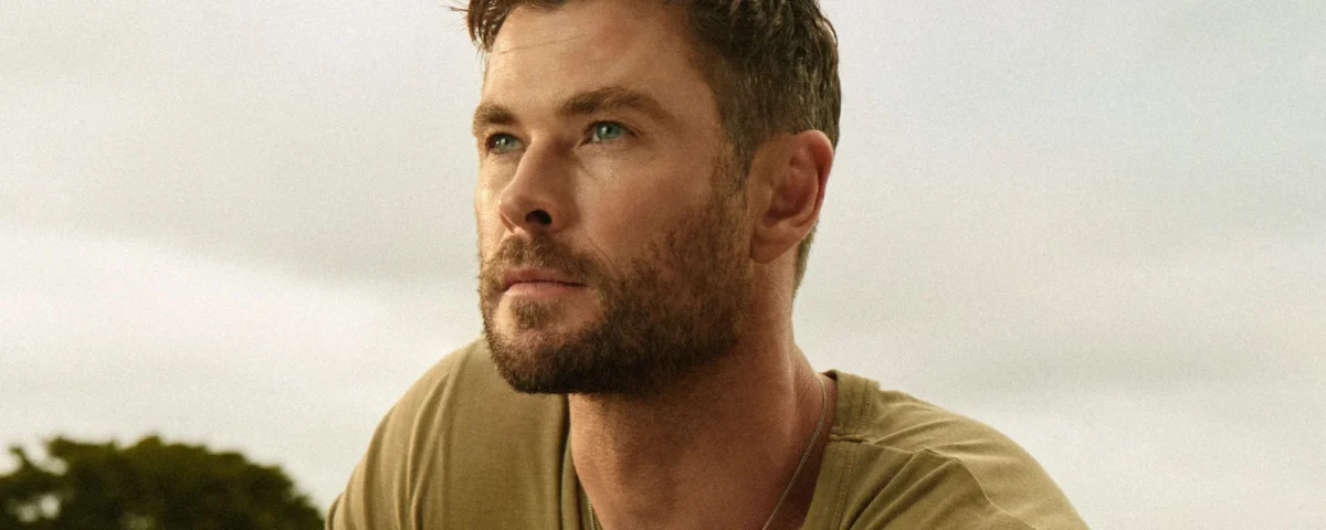 Un homme aux cheveux bruns courts et à la barbe taillée regarde au loin en plein air, vêtu d'un t-shirt kaki et d'un collier - son style rappelle celui de Chris Hemsworth dans Limitless de Disney+. Des arbres et un ciel nuageux sont à l'arrière-plan.