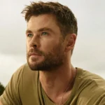 Un homme aux cheveux bruns courts et à la barbe taillée regarde au loin en plein air, vêtu d'un t-shirt kaki et d'un collier - son style rappelle celui de Chris Hemsworth dans Limitless de Disney+. Des arbres et un ciel nuageux sont à l'arrière-plan.