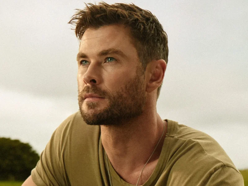 Un homme aux cheveux bruns courts et à la barbe taillée regarde au loin en plein air, vêtu d'un t-shirt kaki et d'un collier - son style rappelle celui de Chris Hemsworth dans Limitless de Disney+. Des arbres et un ciel nuageux sont à l'arrière-plan.