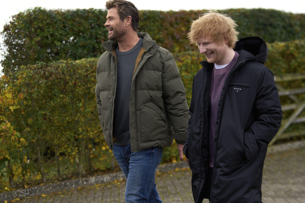 Deux hommes, vêtus de manteaux d'hiver, se promènent en plein air le long d'une haie et discutent amicalement. La scène évoque l'esprit d'aventure que l'on retrouve dans les émissions de Disney+ telles que Limitless, avec Chris Hemsworth.