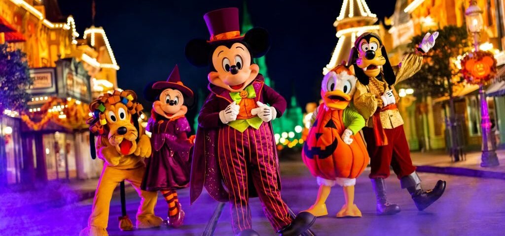 Cinq personnages de Disney en costumes d'Halloween se tiennent dans une rue décorée la nuit pendant Disneyland Halloween 2025, avec du brouillard au sol et des lumières festives en arrière-plan.