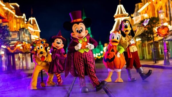 Cinq personnages de Disney en costumes d'Halloween se tiennent dans une rue décorée la nuit pendant Disneyland Halloween 2025, avec du brouillard au sol et des lumières festives en arrière-plan.