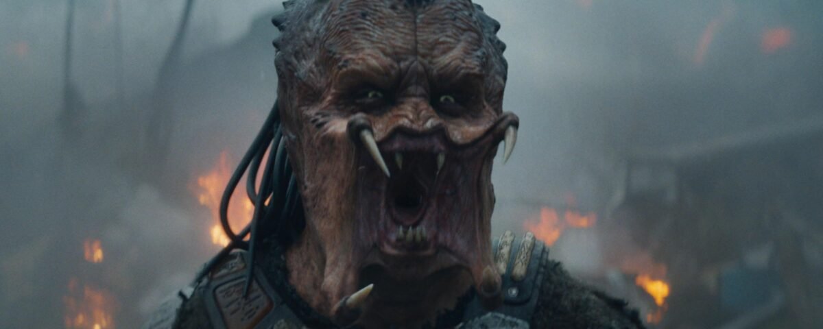 Une créature extraterrestre aux cheveux semblables à des dreadlocks, à la bouche ouverte et aux crocs proéminents se tient devant un arrière-plan enfumé et ardent dans Predator Badlands, le nouvel opus 2025 dont la date de sortie est très attendue.