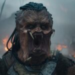 Une créature extraterrestre aux cheveux semblables à des dreadlocks, à la bouche ouverte et aux crocs proéminents se tient devant un arrière-plan enfumé et ardent dans Predator Badlands, le nouvel opus 2025 dont la date de sortie est très attendue.
