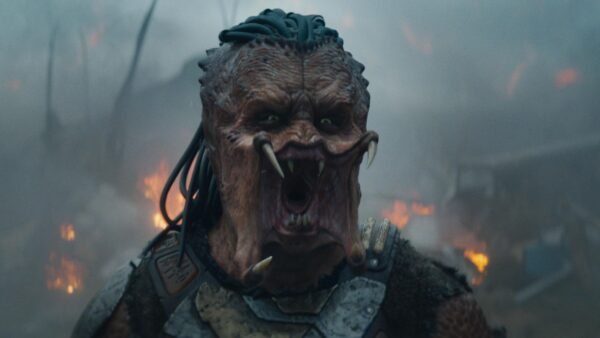 Une créature extraterrestre aux cheveux semblables à des dreadlocks, à la bouche ouverte et aux crocs proéminents se tient devant un arrière-plan enfumé et ardent dans Predator Badlands, le nouvel opus 2025 dont la date de sortie est très attendue.