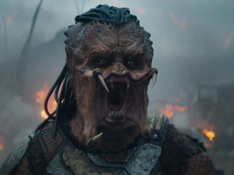 Une créature extraterrestre aux cheveux semblables à des dreadlocks, à la bouche ouverte et aux crocs proéminents se tient devant un arrière-plan enfumé et ardent dans Predator Badlands, le nouvel opus 2025 dont la date de sortie est très attendue.