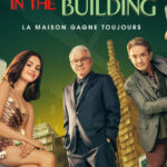 Trois personnes en tenue de soirée posent parmi des cartes à jouer et des dés surdimensionnés, avec de grands immeubles en arrière-plan et le texte "IN THE BUILDING" au-dessus d'eux - une scène de Only Murders In The Building Saison 5, disponible sur Disney+.