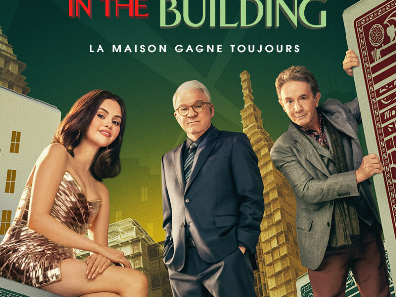 Trois personnes en tenue de soirée posent parmi des cartes à jouer et des dés surdimensionnés, avec de grands immeubles en arrière-plan et le texte "IN THE BUILDING" au-dessus d'eux - une scène de Only Murders In The Building Saison 5, disponible sur Disney+.
