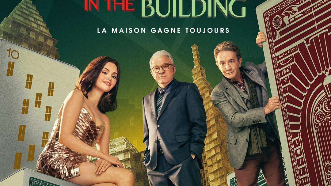 Trois personnes en tenue de soirée posent parmi des cartes à jouer et des dés surdimensionnés, avec de grands immeubles en arrière-plan et le texte "IN THE BUILDING" au-dessus d'eux - une scène de Only Murders In The Building Saison 5, disponible sur Disney+.