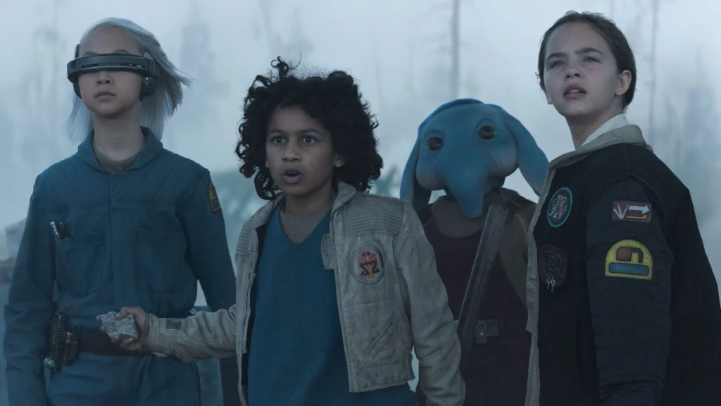 Quatre enfants portant des vêtements futuristes se tiennent ensemble à l'extérieur ; l'un d'eux porte une visière, un autre tient un rocher, et un extraterrestre à la peau bleue et à la trompe se tient derrière eux dans cette scène de la série Disney+ Star Wars Skeleton Crew.