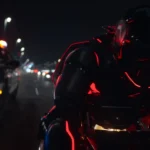 Une personne vêtue d'une combinaison noire futuriste avec des lumières rouges conduit une moto la nuit, poursuivie par une voiture de police dans une rue de la ville - évoquant les scènes de poursuite high-tech vues dans Tron : Ares de Disney avant sa date de sortie.