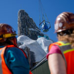 Deux ouvriers du bâtiment observent le levage d'un grand objet par une grue près de la structure rocheuse et enneigée du Palais de Glace Elsa dans le monde de Frozen, sous un ciel bleu limpide.