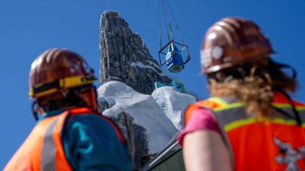 Deux ouvriers du bâtiment observent le levage d'un grand objet par une grue près de la structure rocheuse et enneigée du Palais de Glace Elsa dans le monde de Frozen, sous un ciel bleu limpide.