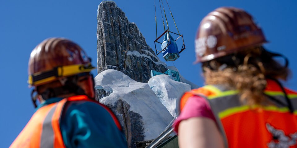 Deux ouvriers du bâtiment observent le levage d'un grand objet par une grue près de la structure rocheuse et enneigée du Palais de Glace Elsa dans le monde de Frozen, sous un ciel bleu limpide.