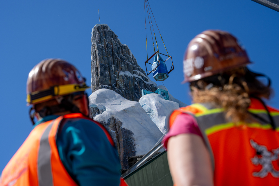 Deux ouvriers du bâtiment observent le levage d'un grand objet par une grue près de la structure rocheuse et enneigée du Palais de Glace Elsa dans le monde de Frozen, sous un ciel bleu limpide.