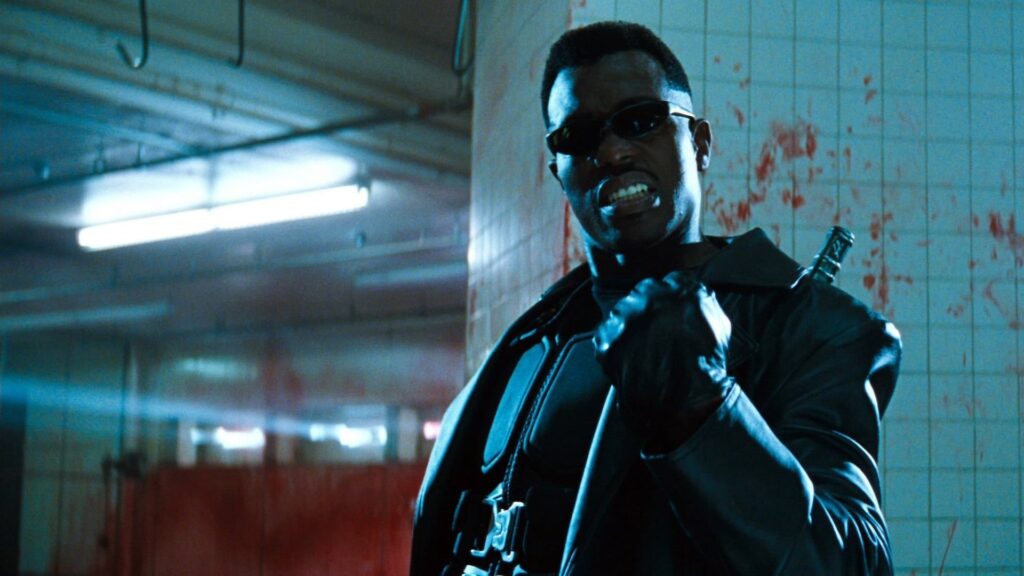Un homme ressemblant au Blade de Marvel, vêtu de cuir noir et portant des lunettes de soleil, se tient dans une pièce éclaboussée de sang, tenant la poignée de l'épée dans son dos comme le personnage emblématique de Mahershala Ali.