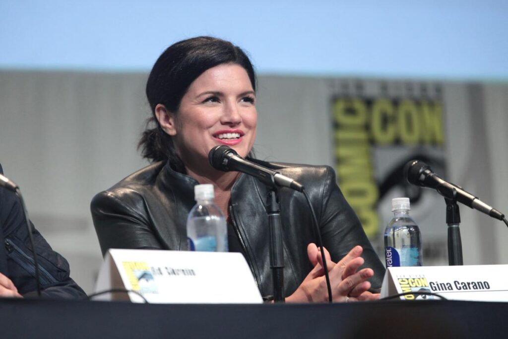Une femme vêtue d'une veste noire, Gina Carano, parle dans un microphone lors d'une table ronde, assise derrière un carton portant son nom et des bouteilles d'eau posées sur la table - probablement pour discuter de son travail avec Disney.