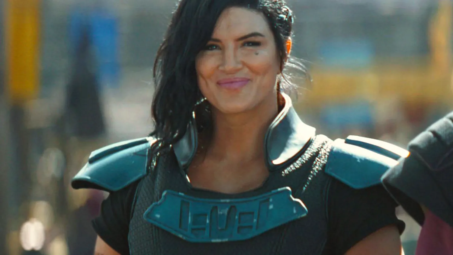 Personne vêtue d'une armure futuriste avec des épaulettes, cheveux noirs coiffés sur le côté, debout à l'extérieur et souriante, rappelant le personnage emblématique de Gina Carano chez Disney.