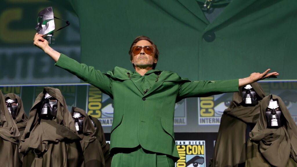 Un homme vêtu d'un costume vert et les bras tendus se tient sur scène avec un masque, entouré de personnes portant des manteaux à capuche au Comic-Con, évoquant une ambiance MCU et faisant allusion au prochain choc de l'Avengers Doomsday.