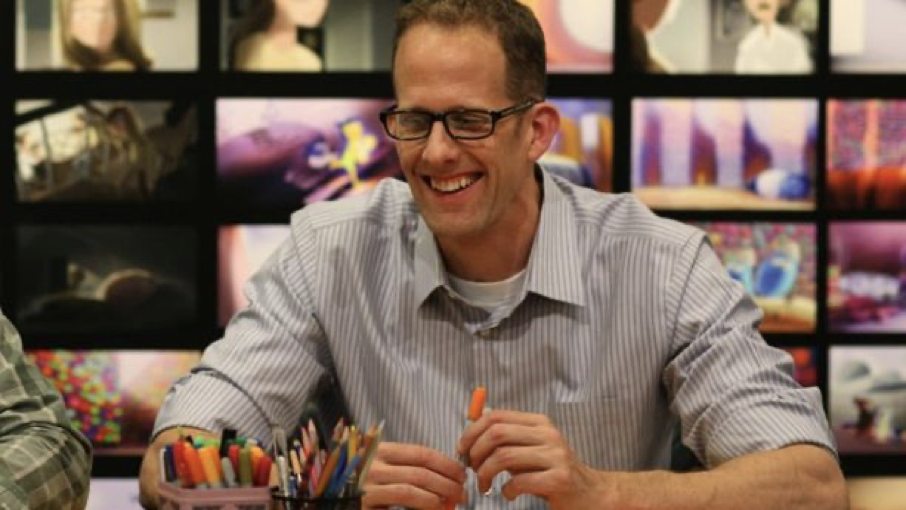 Un homme portant des lunettes et une chemise rayée est assis à une table avec des stylos de couleur, souriant, avec un mur d'images animées affiché derrière lui - et vice-versa, la toile de fond animée semble refléter son énergie joyeuse.