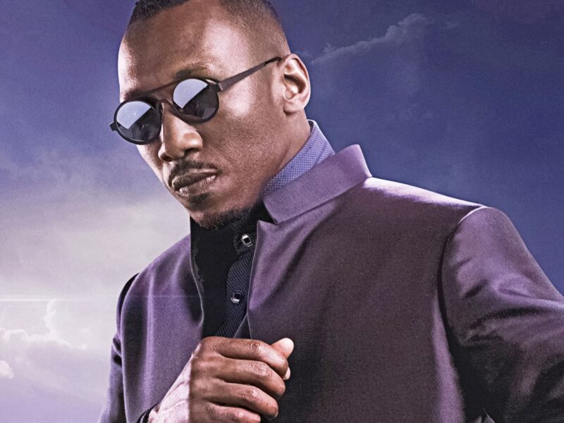 Un homme portant des lunettes de soleil et une veste sombre se tient sur un fond de ciel nuageux. Il incarne la présence confiante du Blade de Marvel, rappelant le style emblématique de Mahershala Ali.