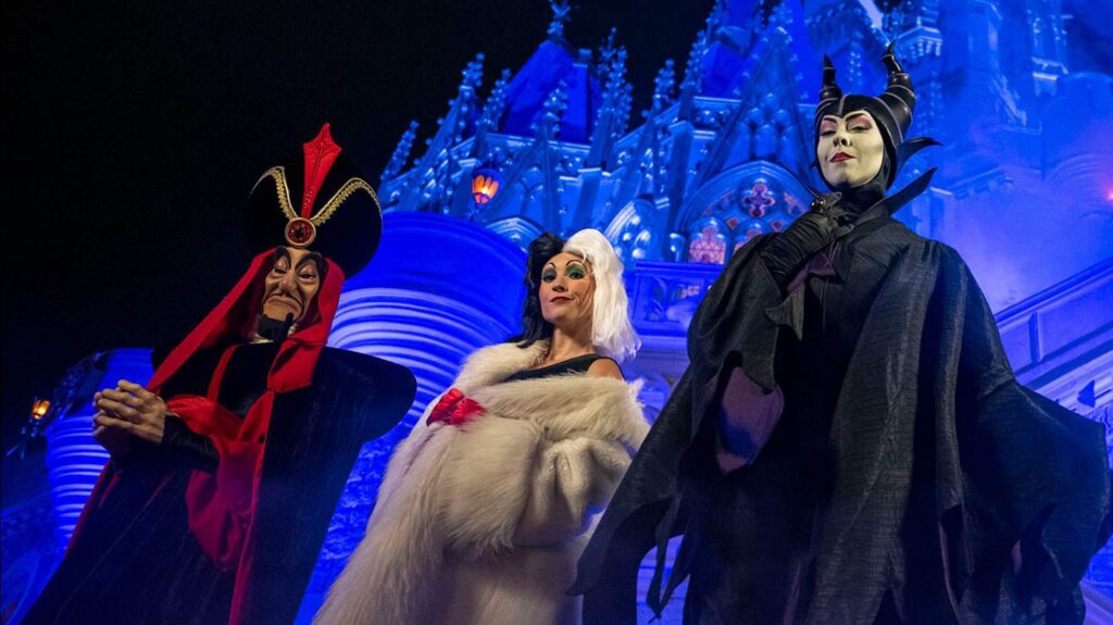 Trois personnes déguisées en Jafar, Cruella de Vil et Maléfique posent devant un château illuminé de lumières bleues lors de la fête d'Halloween 2025 à Disneyland.