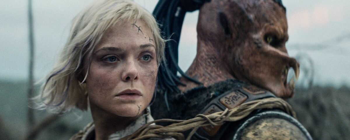 Une femme aux cheveux blonds courts et aux cicatrices faciales se tient dos à dos avec une créature humanoïde portant une armure et un masque, en plein air - une scène saisissante qui rappelle le casting plein d'action de Predator Badlands.