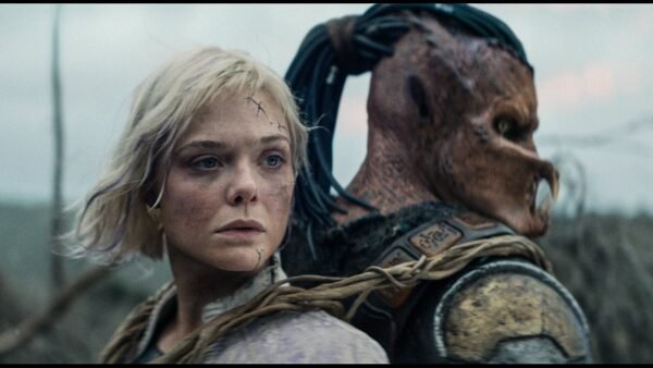 Une femme aux cheveux blonds courts et aux cicatrices faciales se tient dos à dos avec une créature humanoïde portant une armure et un masque, en plein air - une scène saisissante qui rappelle le casting plein d'action de Predator Badlands.