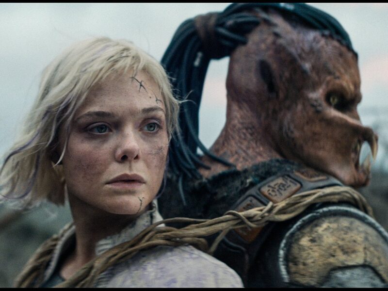 Une femme aux cheveux blonds courts et aux cicatrices faciales se tient dos à dos avec une créature humanoïde portant une armure et un masque, en plein air - une scène saisissante qui rappelle le casting plein d'action de Predator Badlands.