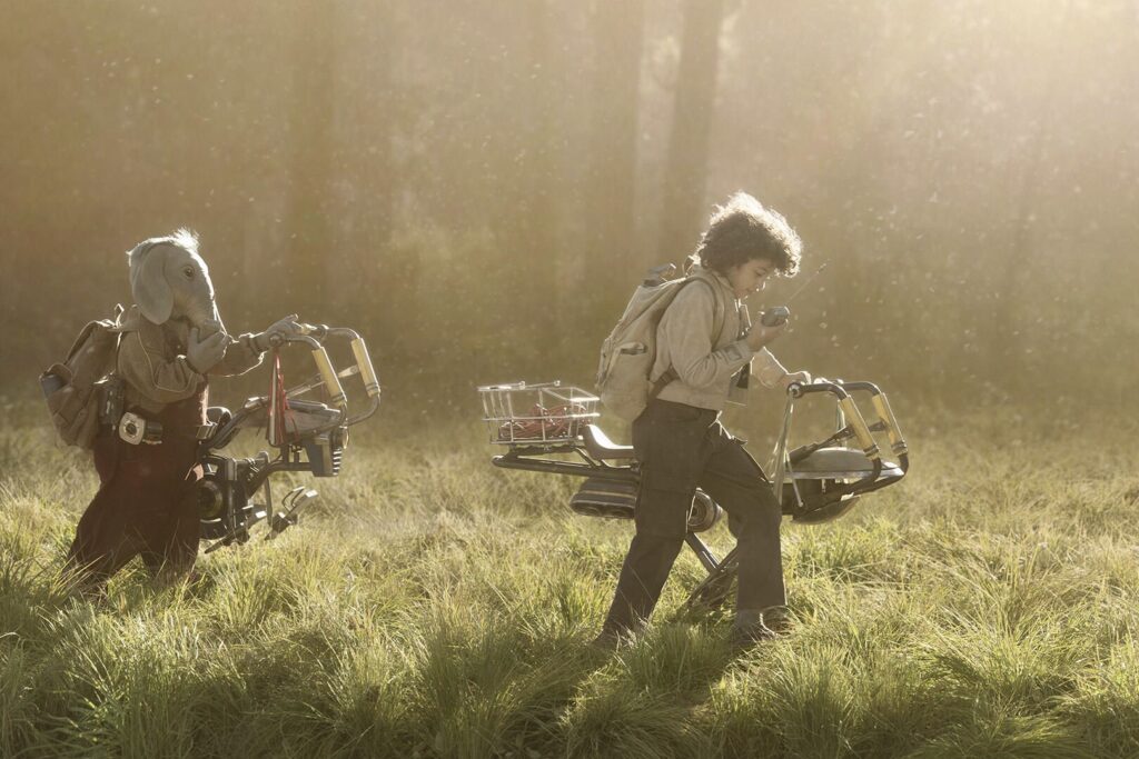 Deux enfants marchent dans les hautes herbes - l'un avec un masque d'âne, l'autre avec des sacs à dos et des cadres de vélo - dans une forêt ensoleillée et brumeuse, rappelant une scène aventureuse de Star Wars Skeleton Crew, bientôt en streaming sur Disney+.