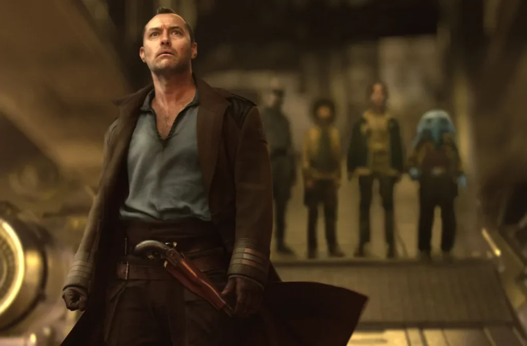 Un homme vêtu d'un long manteau se tient au premier plan et quatre personnes, dont une avec un casque bleu, se tiennent dans l'arrière-plan flou sur le plateau futuriste de Star Wars Skeleton Crew Finale Saison 1 sur Disney+.