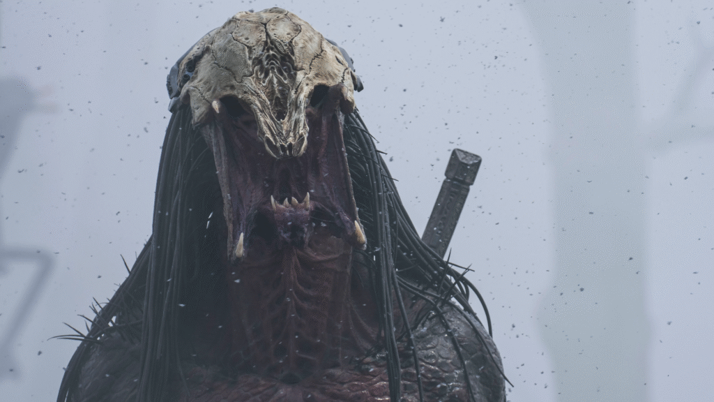 Une créature monstrueuse avec une tête de crâne osseuse, de longues dreads noires et une bouche ouverte se tient dans un environnement brumeux avec une épée attachée dans le dos, évoquant l'aura menaçante de Predator Badlands.