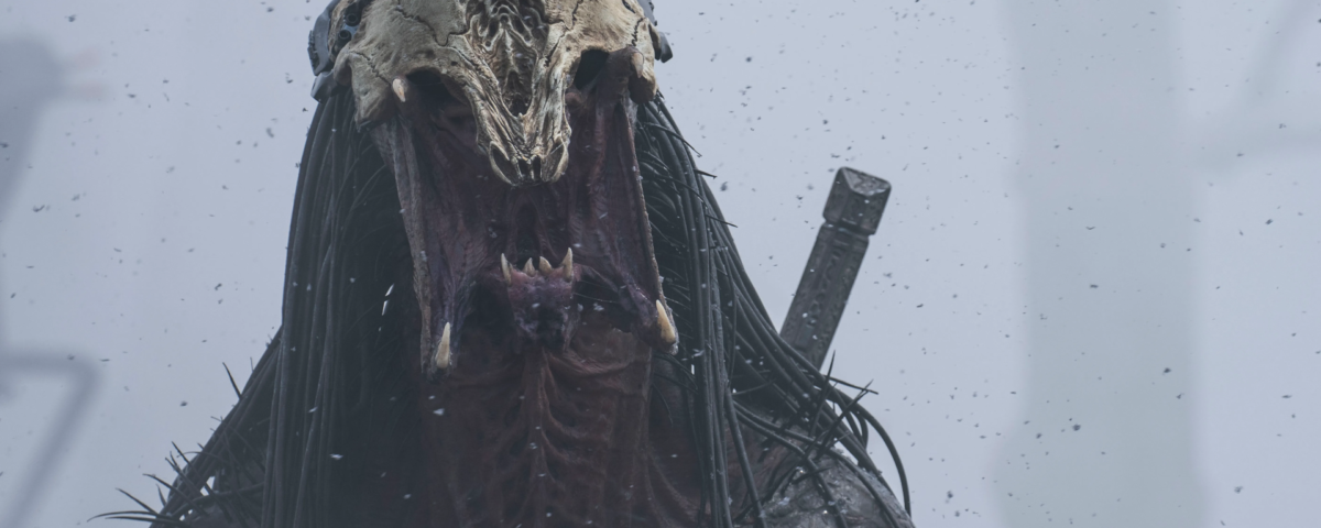 Une créature monstrueuse avec une tête de crâne osseuse, de longues dreads noires et une bouche ouverte se tient dans un environnement brumeux avec une épée attachée dans le dos, évoquant l'aura menaçante de Predator Badlands.