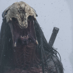 Une créature monstrueuse avec une tête de crâne osseuse, de longues dreads noires et une bouche ouverte se tient dans un environnement brumeux avec une épée attachée dans le dos, évoquant l'aura menaçante de Predator Badlands.