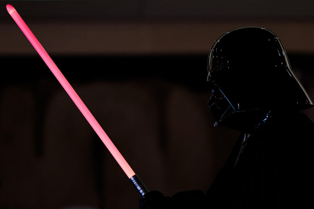 Une personne déguisée en Dark Vador tient un Sabre Laser Dark Vador sur un fond sombre, capturant le look emblématique de Star Wars.