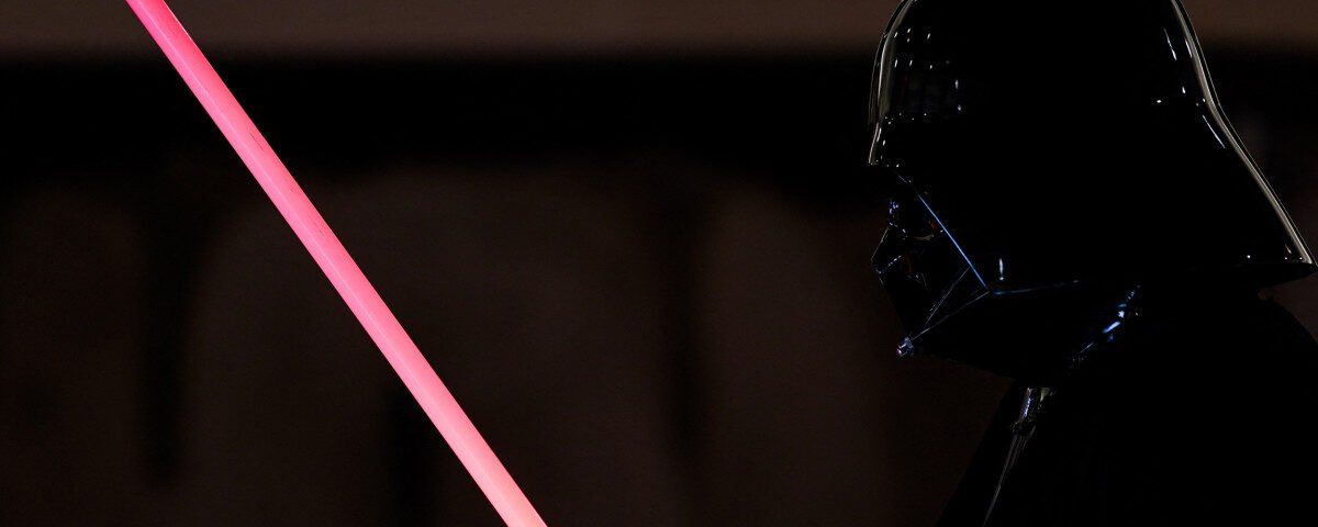 Une personne déguisée en Dark Vador tient un Sabre Laser Dark Vador sur un fond sombre, capturant le look emblématique de Star Wars.