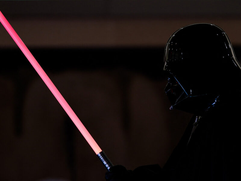 Une personne déguisée en Dark Vador tient un Sabre Laser Dark Vador sur un fond sombre, capturant le look emblématique de Star Wars.