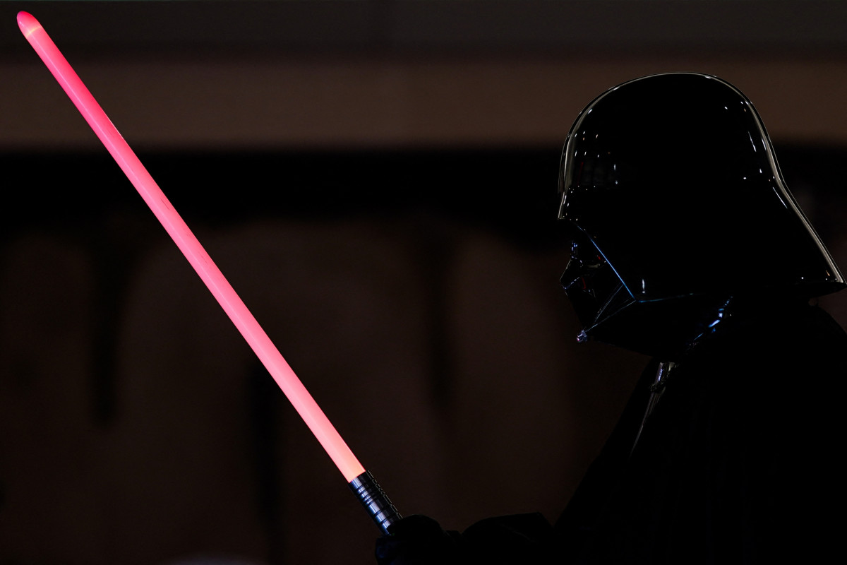 Une personne déguisée en Dark Vador tient un Sabre Laser Dark Vador sur un fond sombre, capturant le look emblématique de Star Wars.