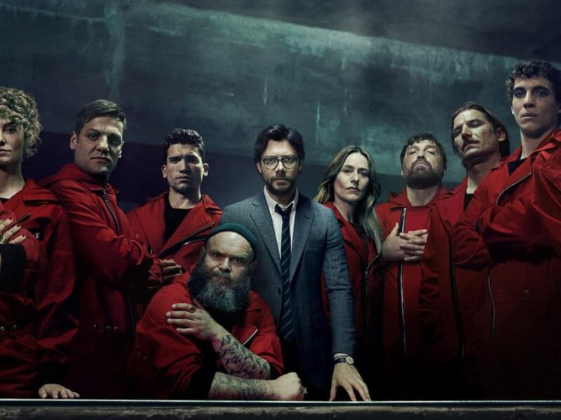 Un groupe de personnes portant des combinaisons rouges et un homme en costume se tiennent ensemble dans une pièce faiblement éclairée, regardant directement la caméra, ce qui rappelle les contenus espagnols populaires disponibles sur Disney+ Espagne.