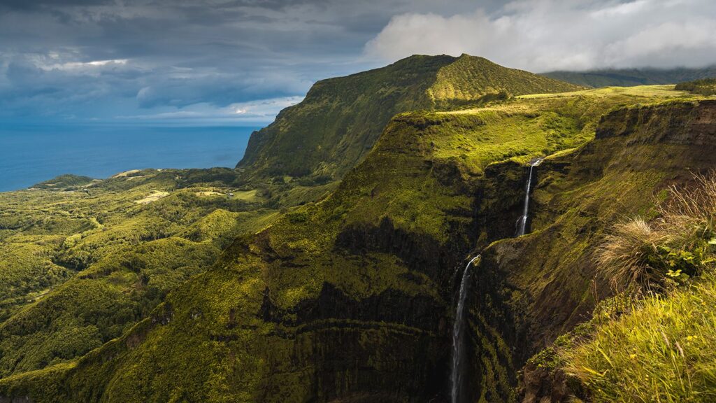 Des falaises verdoyantes avec une haute cascade qui se jette dans une vallée profonde, surplombant l'océan sous un ciel nuageux - une scène digne du National Geographic France.