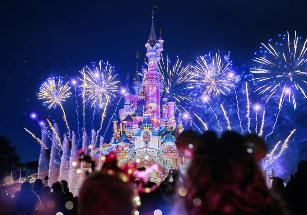 Des feux d'artifice illuminent le ciel nocturne au-dessus d'un château illuminé à Disneyland Paris, avec des silhouettes de spectateurs au premier plan pendant la parade magique de Noël Enchanté Disney.
