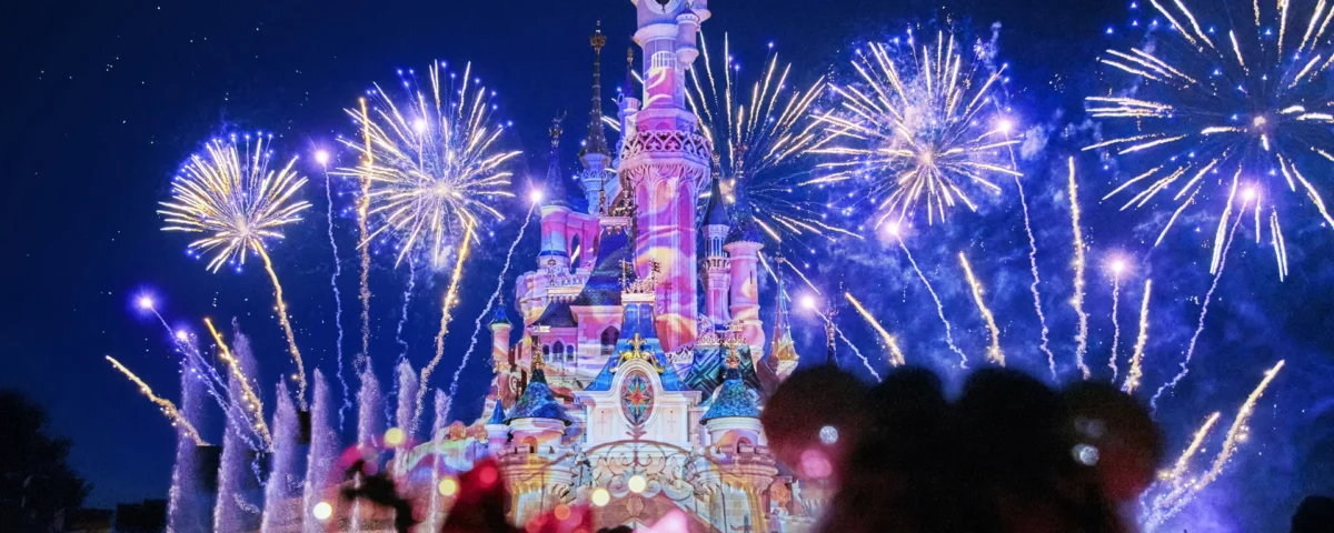 Des feux d'artifice illuminent le ciel nocturne au-dessus d'un château illuminé à Disneyland Paris, avec des silhouettes de spectateurs au premier plan pendant la parade magique de Noël Enchanté Disney.
