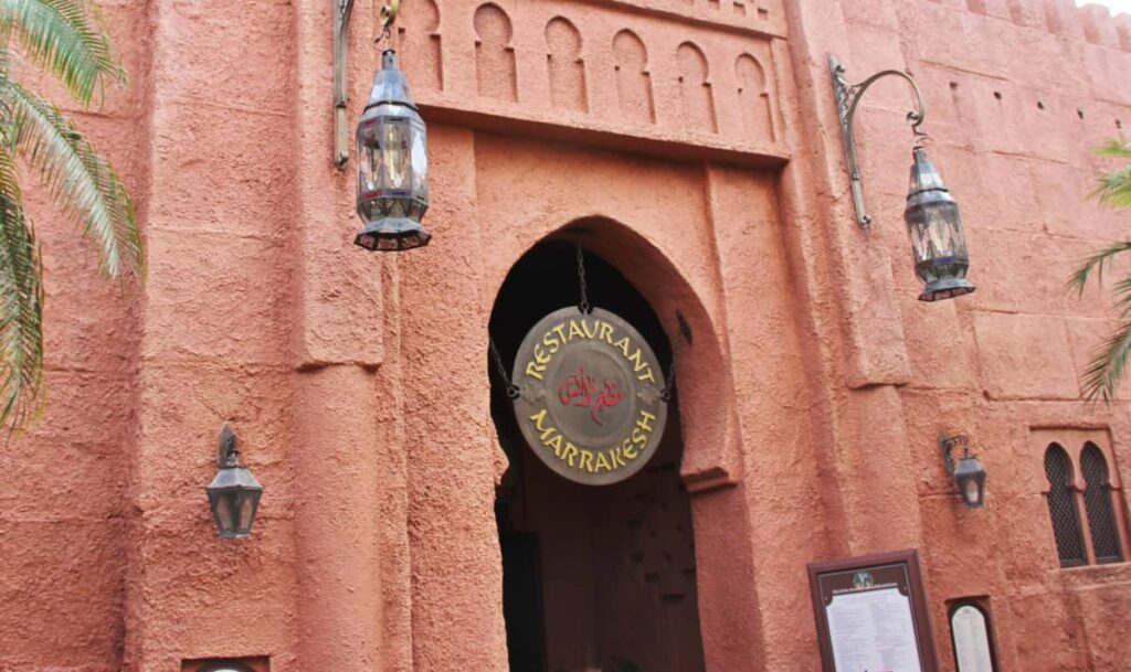 Entrée du restaurant Marrakesh à Disney World Orlando, avec une enseigne ronde suspendue et deux lanternes, sur fond de murs texturés rose-orange et d'arcades ornées dans le pavillon du Maroc.