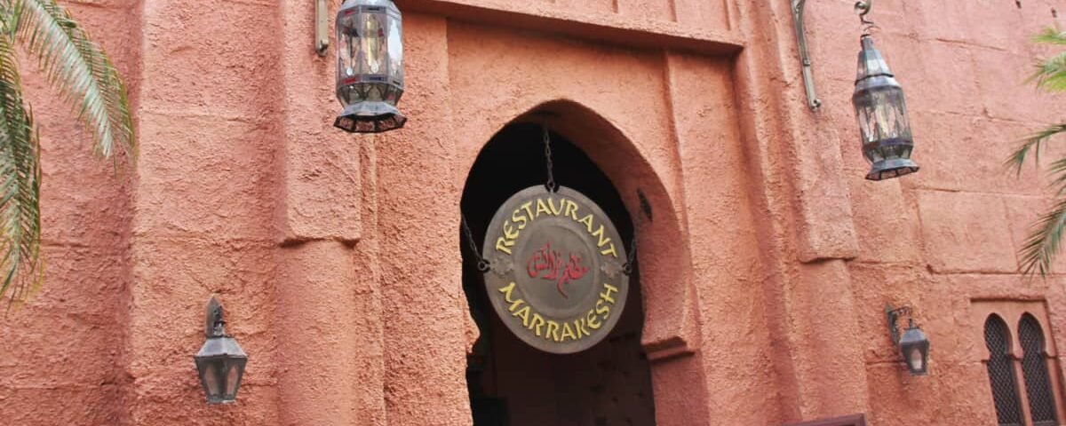 Entrée du restaurant Marrakesh à Disney World Orlando, avec une enseigne ronde suspendue et deux lanternes, sur fond de murs texturés rose-orange et d'arcades ornées dans le pavillon du Maroc.