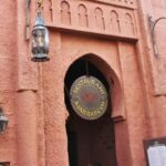 Entrée du restaurant Marrakesh à Disney World Orlando, avec une enseigne ronde suspendue et deux lanternes, sur fond de murs texturés rose-orange et d'arcades ornées dans le pavillon du Maroc.