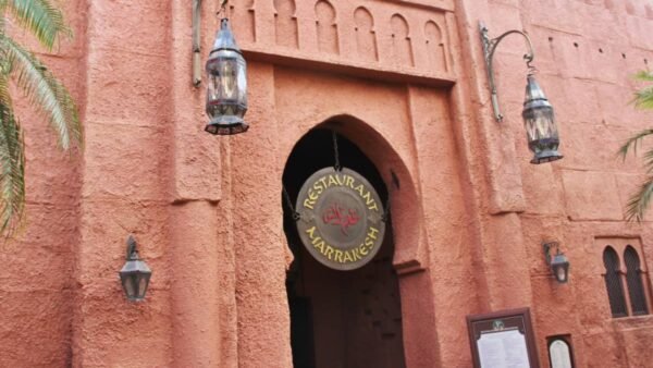 Entrée du restaurant Marrakesh à Disney World Orlando, avec une enseigne ronde suspendue et deux lanternes, sur fond de murs texturés rose-orange et d'arcades ornées dans le pavillon du Maroc.